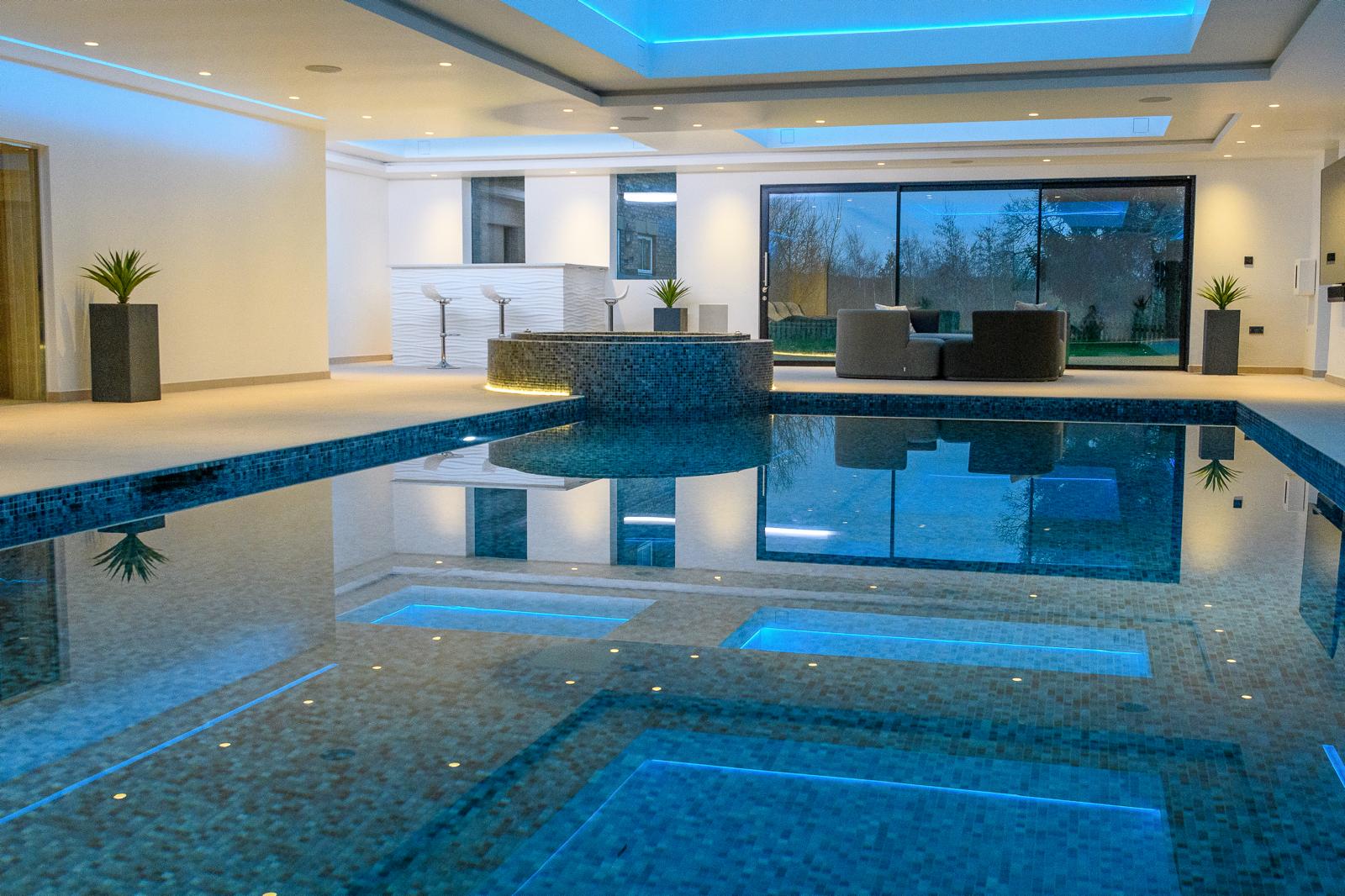 Indoor pool extension lit at night with mosaic tiling, a sunken seating area and full-height glazing